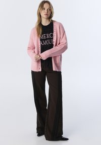 Jasnoróżowy sweter robiony na drutach z guzikami, noszony na czarna strukturalną koszulę, zestawiony z ciemnobrązowymi spodniami z bocznymi paskami.