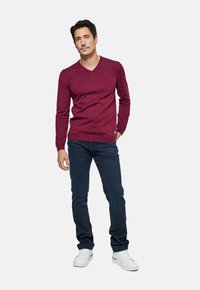 Homme debout portant un pull V en bordeaux, un jean bleu foncé et des baskets blanches devant un fond clair.