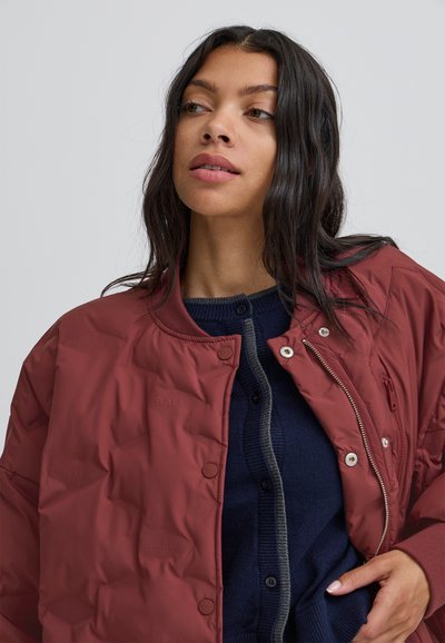 Sarkans quiltots jakas ar noapaļotu apkakli, kas aprīkota ar pogām un rāvējslēdzēju. Valkāta virs tumši zilā kardigāna ar pelēkiem akcentiem.