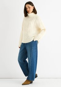 Maglione lavorato a maglia color crema con colletto alto e motivo a treccia, abbinato a jeans blu larghi e mule marroni con tacco largo.