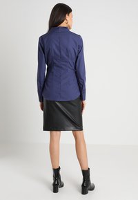 Marineblaues Hemd mit taillierter Passform, langen Ärmeln und Rückennähten, kombiniert mit einem schwarzen, aus Kunstleder gefertigten Bleistiftrock und schwarzen Stiefeletten.