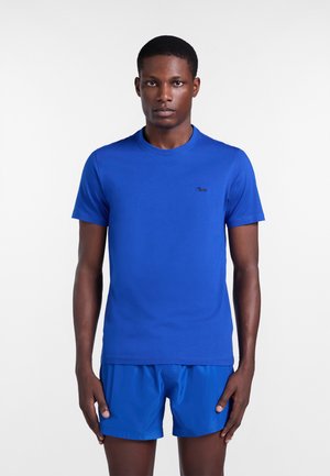 Muž stojaci, oblečený v jednofarebnom jasne modrom tričku s krátkym rukávom a zodpovedajúcich modrých šortkách na bielom pozadí.