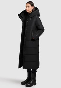 Czarny długi puchowy płaszcz z dużym kapturem, pikowany wzór, kieszenie zapinane na zamek i gładka, błyszcząca faktura. Połączony z czarnymi botkami za kostkę.