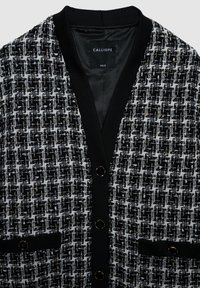 Cardigan noir et blanc avec motif tweed texturé, col et poignets côtelés noirs, boutons dorés en accent, et poches avant.