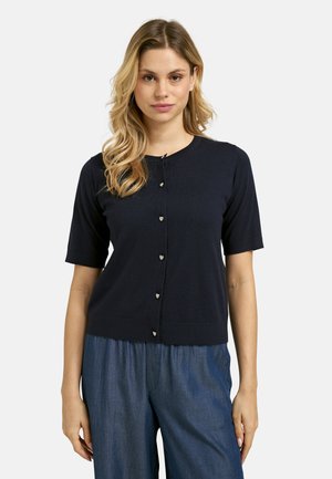 Woman wearing a black short-sleeve buttoned cardigan with heart-shaped buttons and blue loose-fitting pants, standing against a white background.