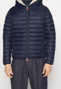 Blå vadderad jacka med huva, med quiltade sektioner, en framtill dragkedja med orange dragflik och ett mjukt fleecfoder.