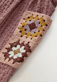 Nærbillede af et tekstureret støvet lilla stof med en ærmepatch, der har flerfarvede hæklede firkanter i brun, sennep, lavendel, hvid og beige.