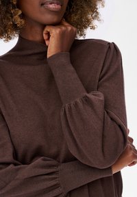 Zbliżenie na osobę w brązowym swetrze z wysokim kołnierzem i bufkami, opierającą podbródek na zaciśniętej pięści.