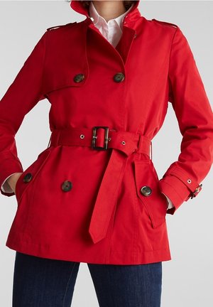 Vrouw die een rode trenchcoat met riem en grote knopen draagt, staand met handen in de zakken, gecombineerd met donkerblauwe jeans en een wit overhemd.