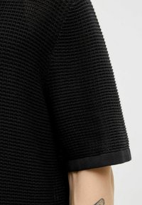 Black textured short-sleeve shirt with a ribbed pattern and a solid black trim at the sleeve hems, featuring a visible tattoo on the arm.