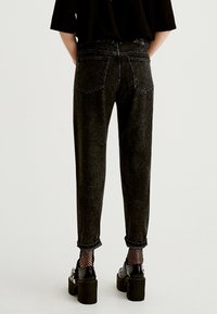 Person som bär uppvikta svarta jeans, fisknätssockor, plattformsskor i svart och en svart topp, står med händerna bakom ryggen.