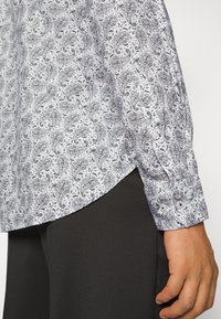 Chemise blanche à manches longues avec un motif cachemire sombre complexe portée avec un pantalon noir, montrant le poignet boutonné et le détail de l'ourlet latéral.