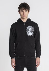 Crna zip-up dukserica od pamuka, s grafičkim printom glave okružene zmijama na lijevoj strani prsnog dijela. Prednji džepovi.