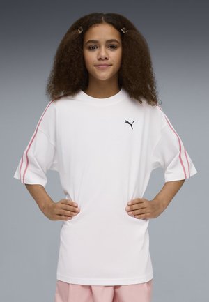 Giovane con capelli ricci che indossa una maglietta bianca oversize Puma con strisce rosa sulle maniche e pantaloni rosa chiaro, con le mani sui fianchi.