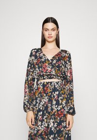 Top floral envolvente com mangas compridas, apresentando um decote V profundo e cintura franzida. Fundo azul-marinho escuro com flores de várias cores e botões.