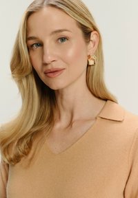 Béžový sveter s výstrihom do V a golierovým dizajnom. Osoba má na sebe štruktúrované zlaté geometrické náušnice. Hladké blond vlasy.