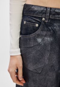 Tamsūs džinsai su tekstūruotu paviršiumi, turintys priekinę ir galinę kišenes, metalinius elementus ir siuvimo detales. Artimas audinio vaizdas.