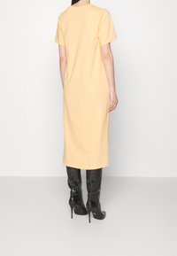 Jasnożółta bawełniana sukienka z krótkim rękawem, prostym krojem i do kolan, noszona z czarnymi błyszczącymi kozakami do kolan.