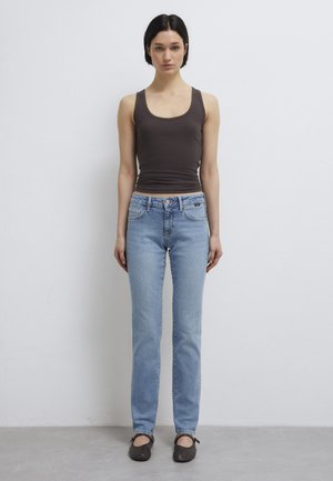 Woman standing against white wall wearing dark brown tank top, light blue straight-leg jeans, and black ballet flats, facing forward.