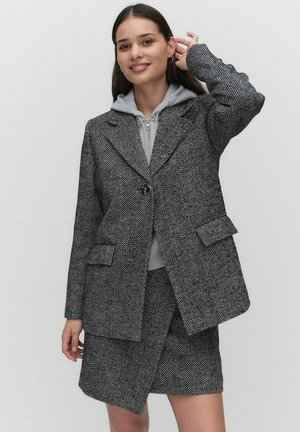 Blazer à motif chevrons noir et blanc, avec revers crantés, deux poches avant et une fermeture à un bouton, superposé à un hoodie gris.