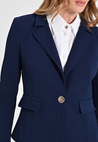 Blazer marine ajusté avec revers crantés, fermeture à un bouton doré, et deux poches angleées. Fabriqué en tissu lisse avec une coupe structurée.