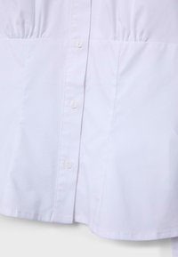 Section de chemise blanche montrant trois petits boutons avec des fronces de tissu au-dessus de la couture de la taille et un ourlet net en dessous.