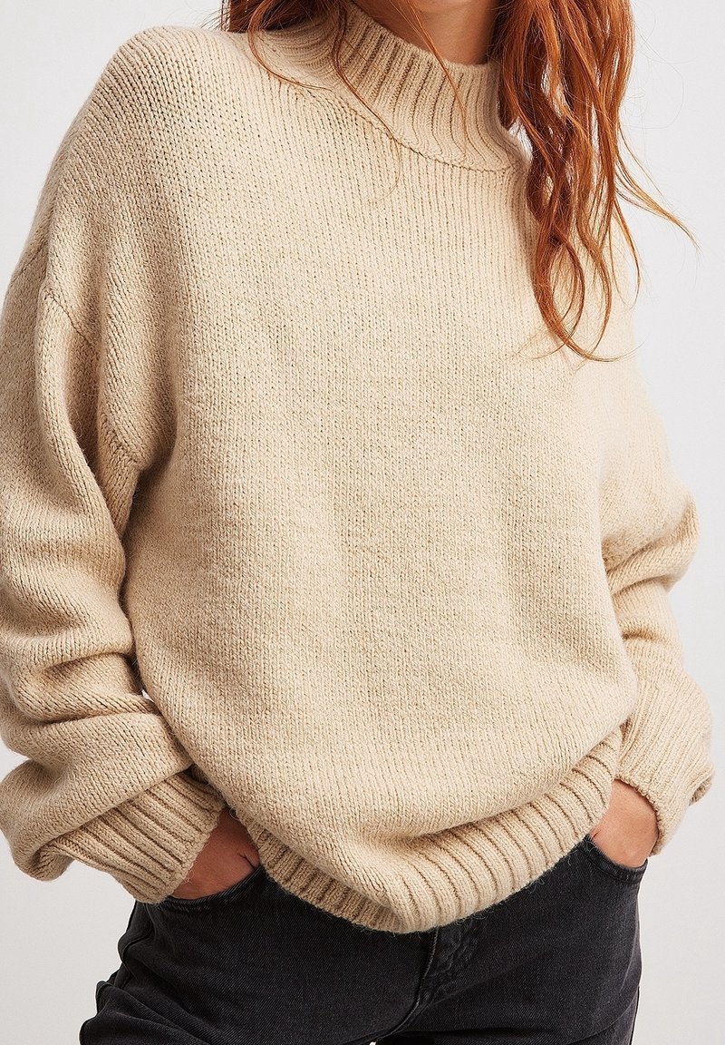 Beżowy sweter dziergany z wysokim prążkowanym kołnierzem, luźnymi rękawami i prążkowanym dołem. Miękka faktura, luźny krój i minimalne zdobienia.