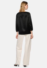 Woman standing, facing away, wearing a loose black satin blouse, white wide-leg pants, black heels, and black earrings on a light background.