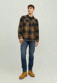 Νεαρός άνδρας που στέκεται, φορώντας καφέ και ναυτική καρό πουκάμισο, μπλε τζιν και καφέ παπούτσια με κορδόνια, μπροστά από έναν απλό ανοιχτό φόντο.