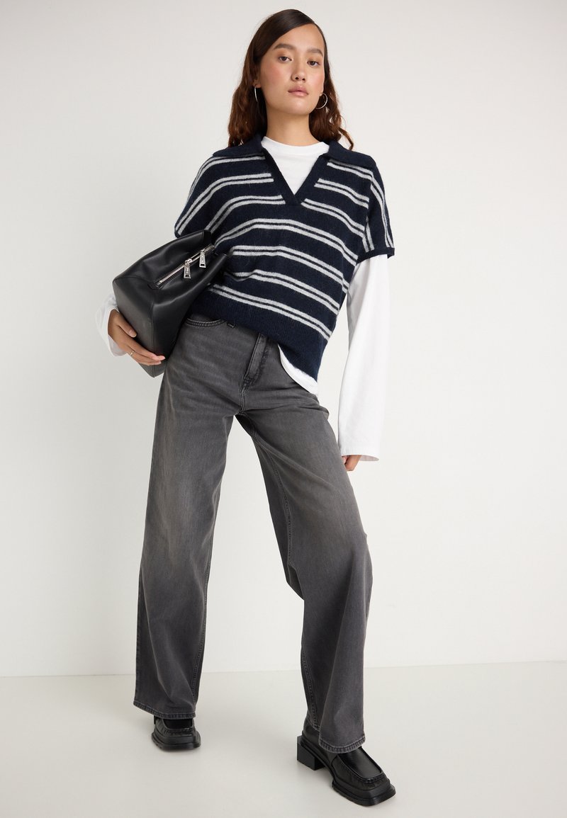 Granatowy i biały, paskowany sweter bez rękawów z dekoltem w serek, nałożony na biały, długi rękaw. Połączony z szarymi spodniami z szerokimi nogawkami i czarnymi, masywnymi butami.