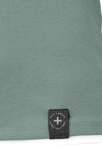 Primer plano de tela verde con estrías con una pequeña etiqueta negra que presenta una cruz blanca y texto circular cerca del dobladillo.