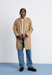 Beige længere frakke med krave, knaplukning og brystlogo, parret med blå denimbukser med et subtilt mønster.
