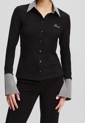 Chemise noire cintrée boutonnée avec col gris contrasté et larges poignets, brodée "Karl" sur la poitrine gauche, portée avec un pantalon noir.