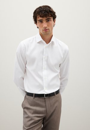 Hombre con cabello rizado, vistiendo una camisa blanca de botones metida dentro de pantalones marrones con un cinturón negro, de pie frente a un fondo simple.