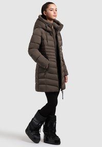 Chaqueta de plumas marrón con cuello alto, cierre de cremallera y capucha ajustable. Leggings negros y botas negras aisladas completan el conjunto.