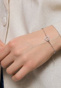 Srebrna bransoletka z wiszącym wisiorkiem w kształcie serca, ozdobionym przezroczystymi cyrkoniami. Wzór łańcuszka, noszona na jasnej skórze dłoni na jasnym tle.