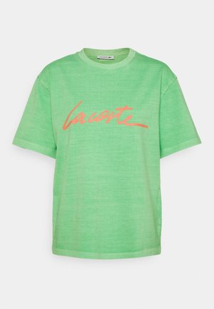 T-shirt vert à manches courtes avec un col rond et un texte stylisé orange "Lacoste" sur la poitrine.
