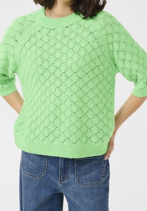 Femme portant un pull en maille vert clair à manches courtes avec un motif festonné et un jean en denim bleu, debout les mains sur les hanches.