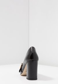 Chaussure à talon haut en cuir noir avec un bout arrondi, un talon bloc haut et un détail de nœud décoratif sur le côté. Texture lisse, design minimaliste.