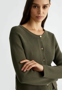 Cardigan a coste verde oliva con collo rotondo, bottoni dorati e maniche lunghe. Il tessuto testurizzato presenta linee verticali.