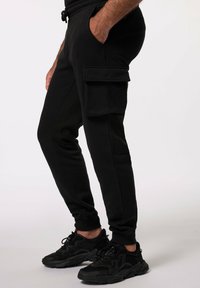 Schwarze Jogginghose mit elastischem Bund, Seitentaschen und einer Cargo-Tasche. Das Material ist weich und strukturiert, kombiniert mit schwarzen Turnschuhen.