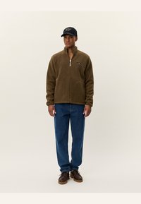 Brown fleece jacket with zip, stand collar, and front pockets. Worn with blue jeans and brown shoes, complemented by a black cap.