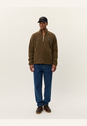 Brown fleece jacket with zip, stand collar, and front pockets. Worn with blue jeans and brown shoes, complemented by a black cap.