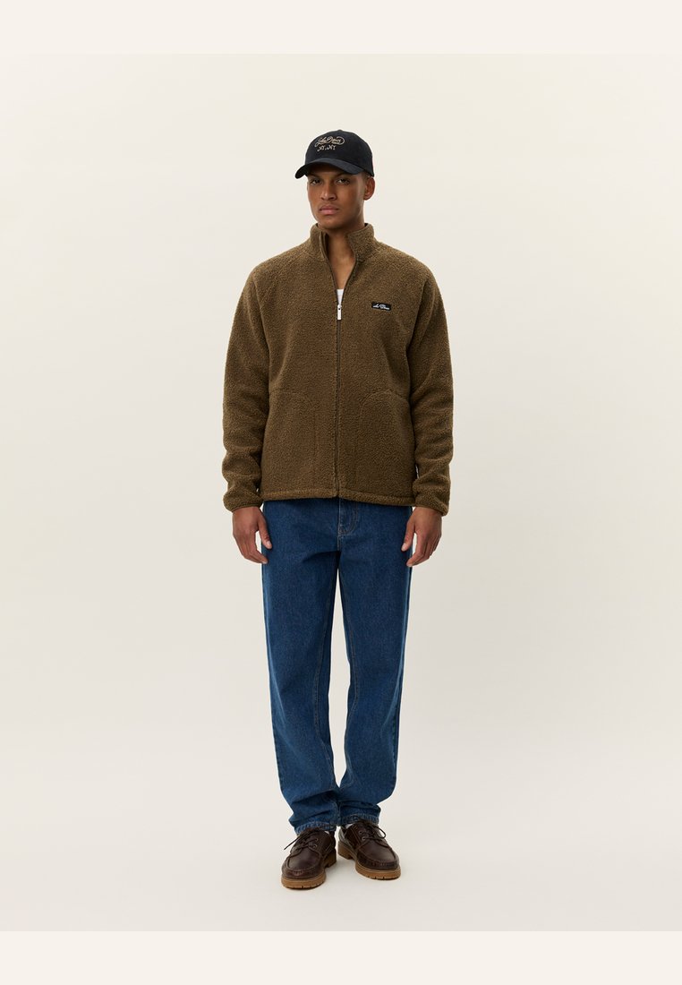 Brown fleece jacket with zip, stand collar, and front pockets. Worn with blue jeans and brown shoes, complemented by a black cap.