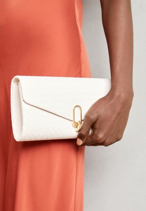 Hand hält eine weiße gewebte Clutch mit goldfarbenem Verschluss vor einem rostfarbenen Stoffhintergrund.