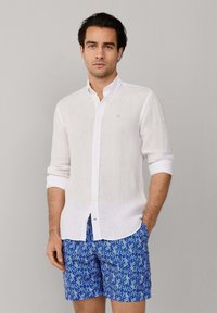 Muž v bielej košeli s dlhými rukávmi vyhrnutými až po lakte a modrých šortkách s vzorom stojí pred jednoduchým sivým pozadím.