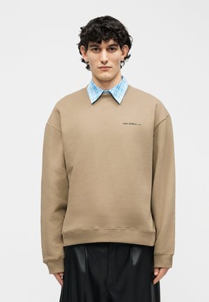 Ung mand med krøllet sort hår iført beige sweatshirt over blåternet skjorte og sorte læderbukser, stående mod hvid baggrund.