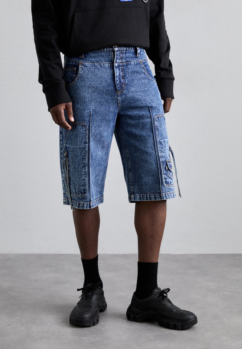 Denim cargo shorts in light blue with contrasting pocket details, zipper closures, and a relaxed fit, paired with black shoes and socks.