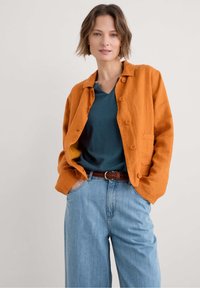 Oranžové lněné sáčko s velkými knoflíky, nošené přes teal tričko s výstřihem do V. Modré široké džíny a hnědý pletený pásek dotvářejí celý outfit.