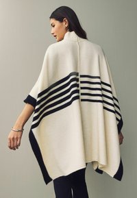 Knit poncho in cream with black striped pattern, high collar, and open sides. Soft texture and relaxed, flowing shape.